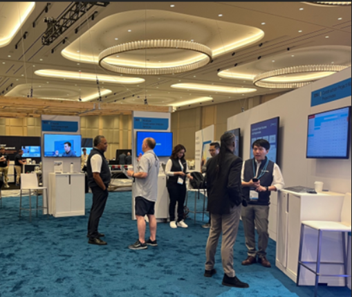 Exhibition booth area at a conference, featuring several people engaging in conversations. The space includes branded counters, mounted screens displaying digital content, and overhead signage. The carpet is blue and the room has modern, circular ceiling lights.