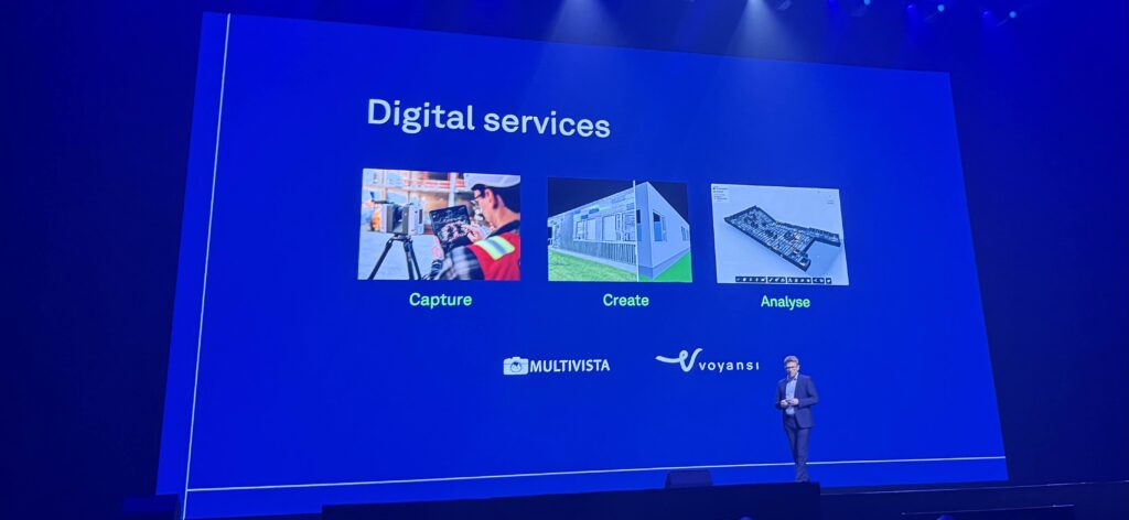 Slide from a keynote presentation titled "Digital services," showing three service pillars: "Capture" with an image of a field technician using a scanner, "Create" with a 3D building model, and "Analyse" with a digital floor plan. Logos of Multivista and Voyansi are shown below the respective pillars, with a speaker standing on stage in front of the slide.